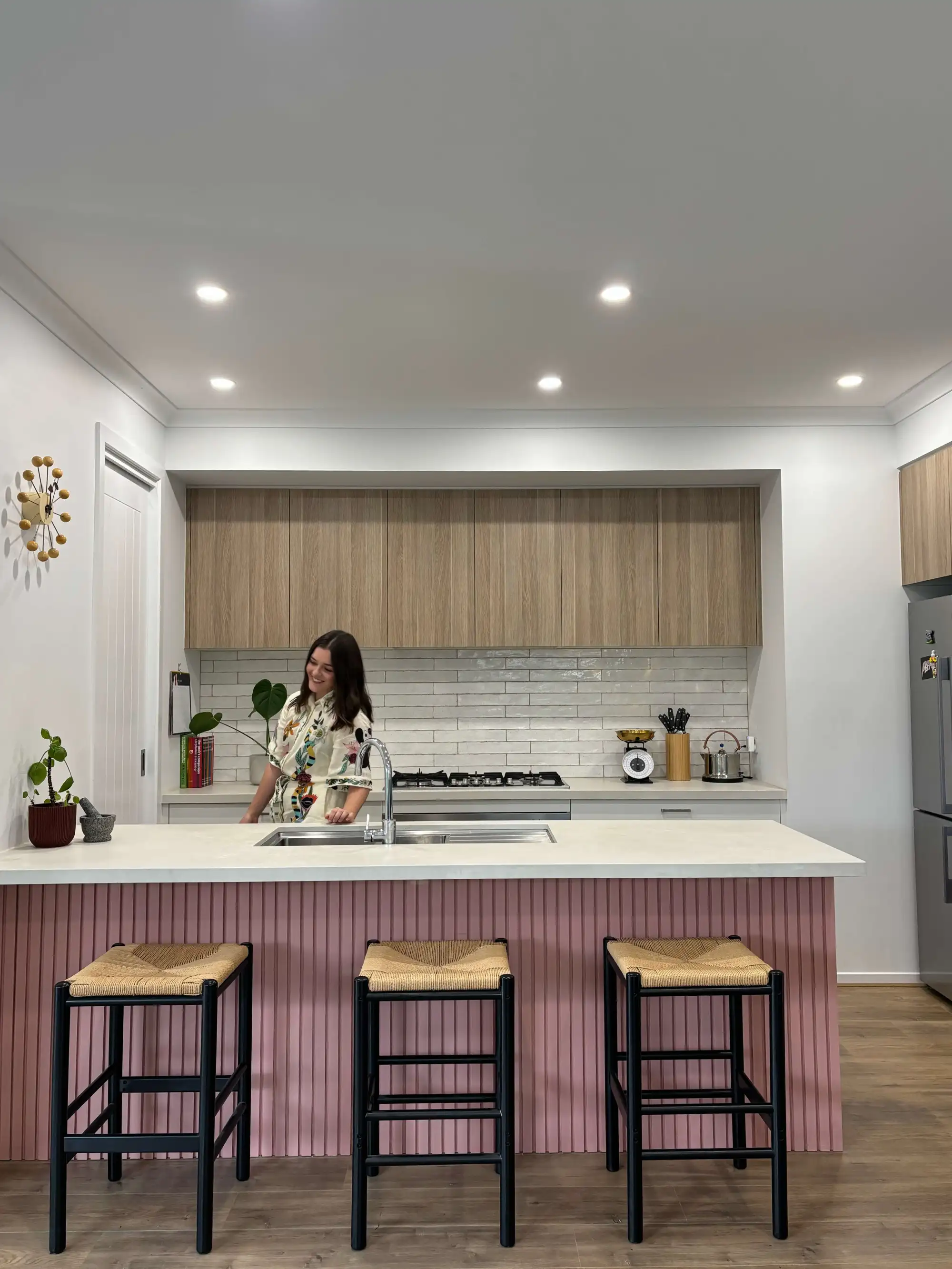 girl standing in kitchen with timber cabinetry, pink benchtop, she has brown hair, and is wearing a floral dress